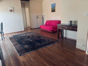 Um quarto com piso de madeira, um tapete grande com padrão, um sofá rosa, uma mesinha e um quadro na parede.