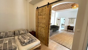 A bedroom with a bed, a nightstand, a wooden sliding door, and a view into a living area with a sofa and a rug. - Danube Atelier – The Garden Apartment (Rossatz-Arnsdorf)