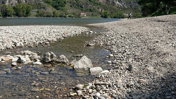 A rocky riverbank with clear water, a mountain in the background, and a person walking. - Danube Atelier – The Garden Apartment (Rossatz-Arnsdorf)