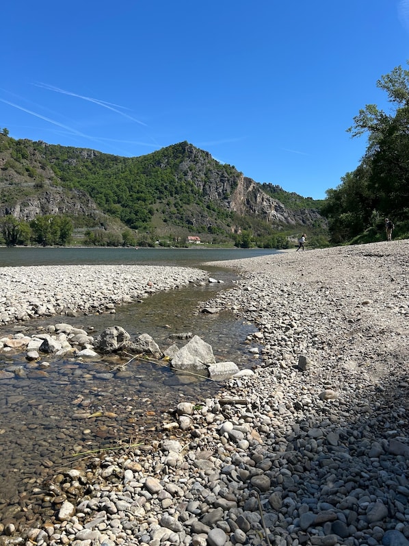 A rocky riverbank with clear water, a distant house, and a mountain in the background.