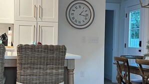 Een keuken met witte kastjes, een wandklok, rieten stoelen en een eethoek met houten stoelen.