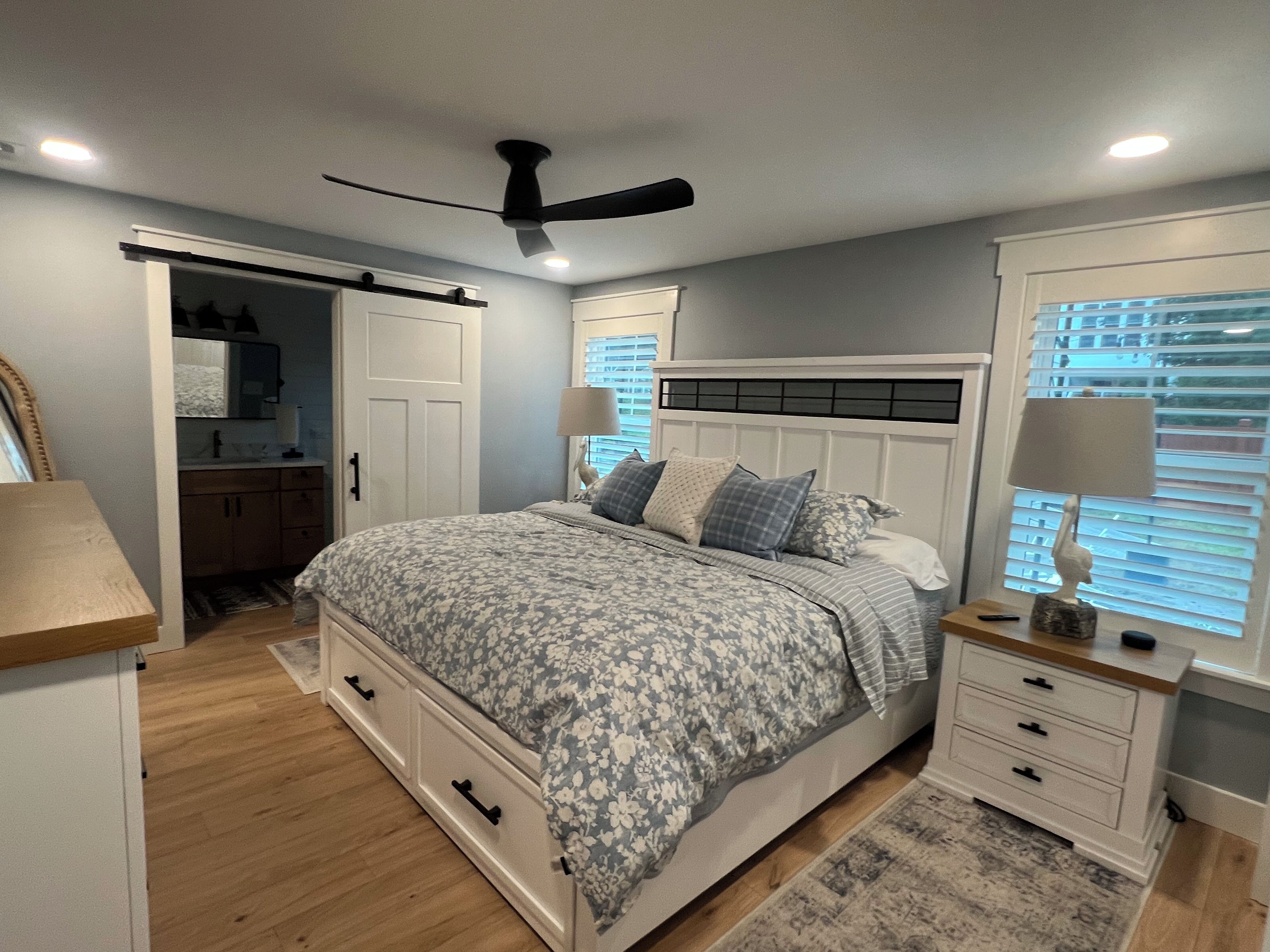 Une chambre à coucher comprenant un lit, des tables de chevet, un ventilateur de plafond, une armoire et une fenêtre avec des stores.