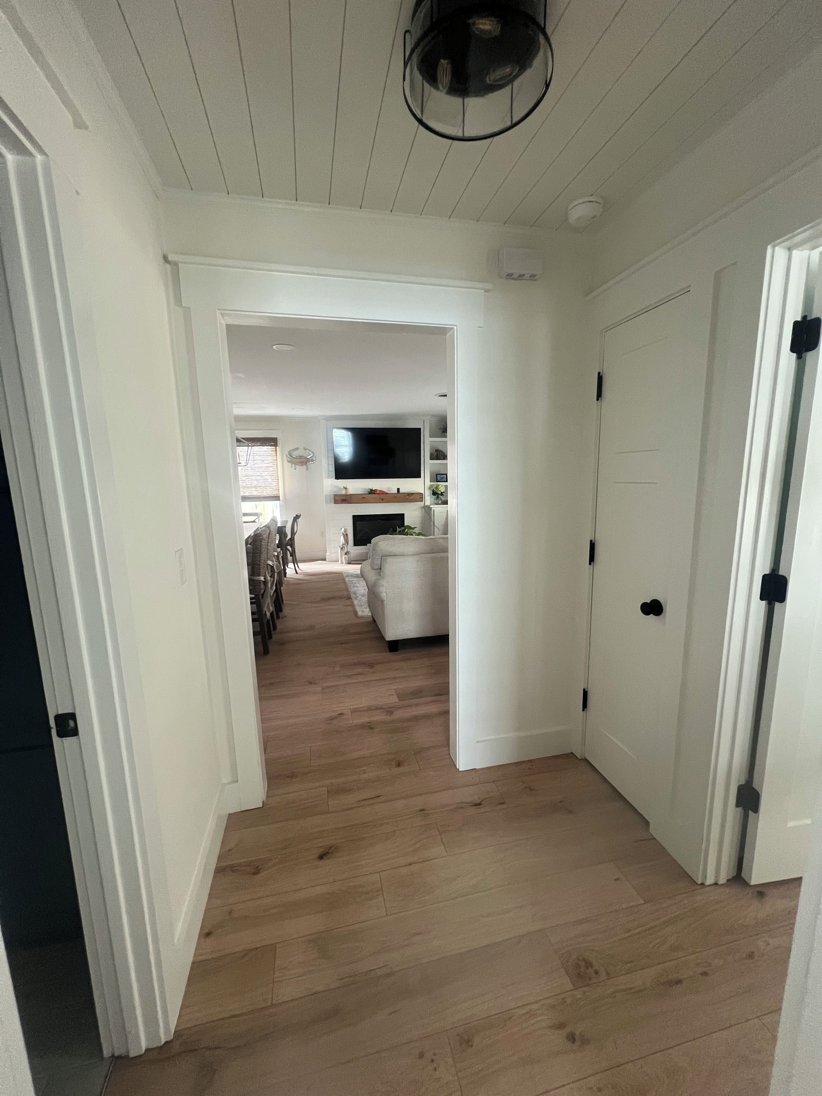 Un couloir au plancher en bois mène à un salon équipé d’un canapé et d’un téléviseur à écran plat fixé au mur.