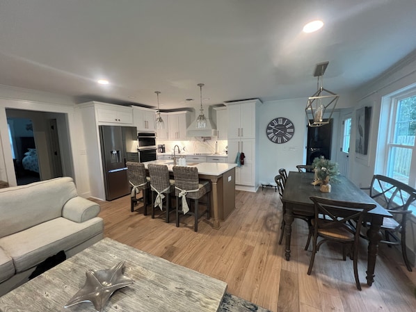 Une cuisine moderne avec des meubles blancs, un coin repas avec une table et des chaises en bois, et un salon avec un canapé et une table basse.