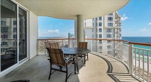 A balcony with a round table and chairs, a view of the ocean, and a building with multiple balconies. - Navarre Beach Colony Gulf Front  / West 15B w/chair service (Navarre)