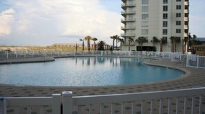 A swimming pool with a surrounding deck, palm trees, and a high-rise building in the background. - Navarre Beach Colony Gulf Front  / West 15B w/chair service (Navarre)