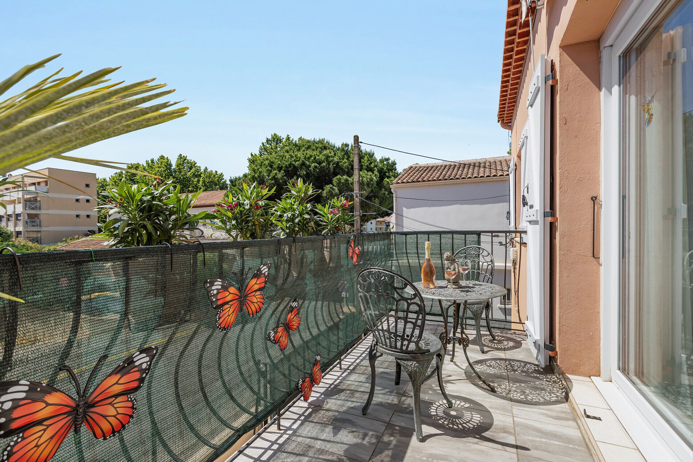 A balcony with a table and chairs, a green mesh fence with butterfly designs, and a building in the background.