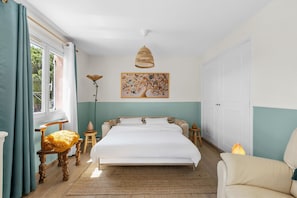 Ein Schlafzimmer mit einem Bett, einem Nachttisch, einer Lampe, einem Sessel, einem Fenster mit Vorhängen und einem Bild an der Wand.