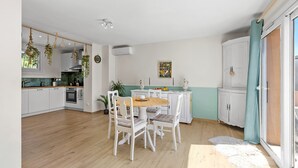 Une cuisine avec des meubles blancs, une table à manger avec des chaises, des plantes suspendues et un tableau au mur.