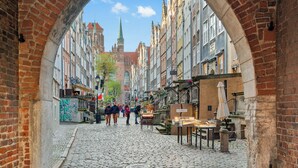 A cobblestone street with brick buildings, a church spire, and outdoor seating areas. - Leśna Aura by Grand Apartments (Sopot)