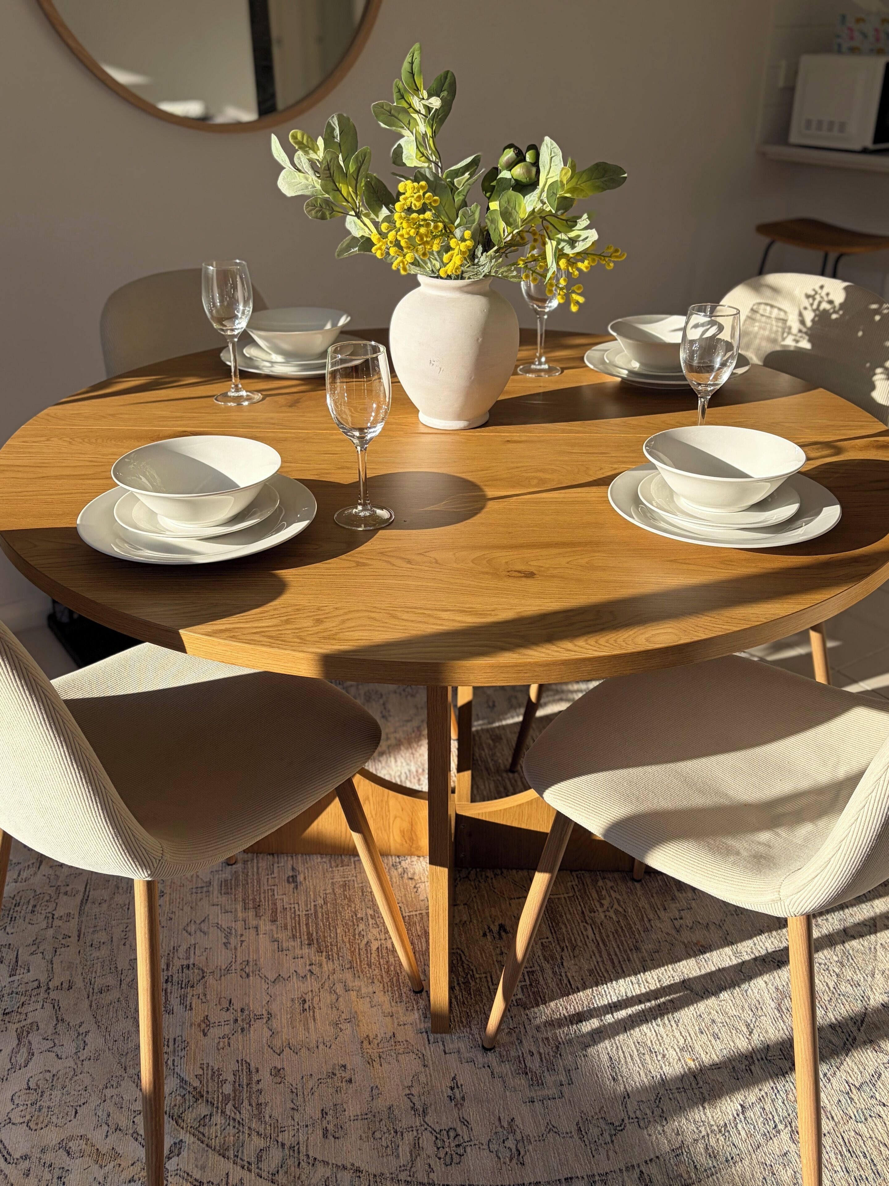 Une table de salle à manger ronde en bois, avec de l’argenterie blanche et des verres, entourée de chaises de couleur beige, et un vase rempli de fleurs jaunes.