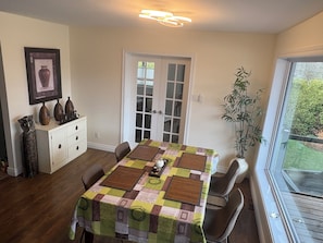 A dining area with a table covered in a patchwork tablecloth, a white cabinet with decorative items, a door with glass panels, and a potted plant by a window.