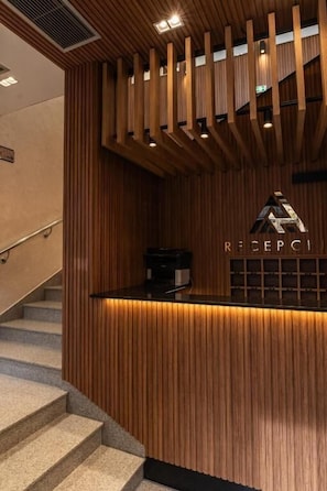 A reception desk with a wooden finish, a staircase to the left, and a sign that reads 'RECEPTION'.