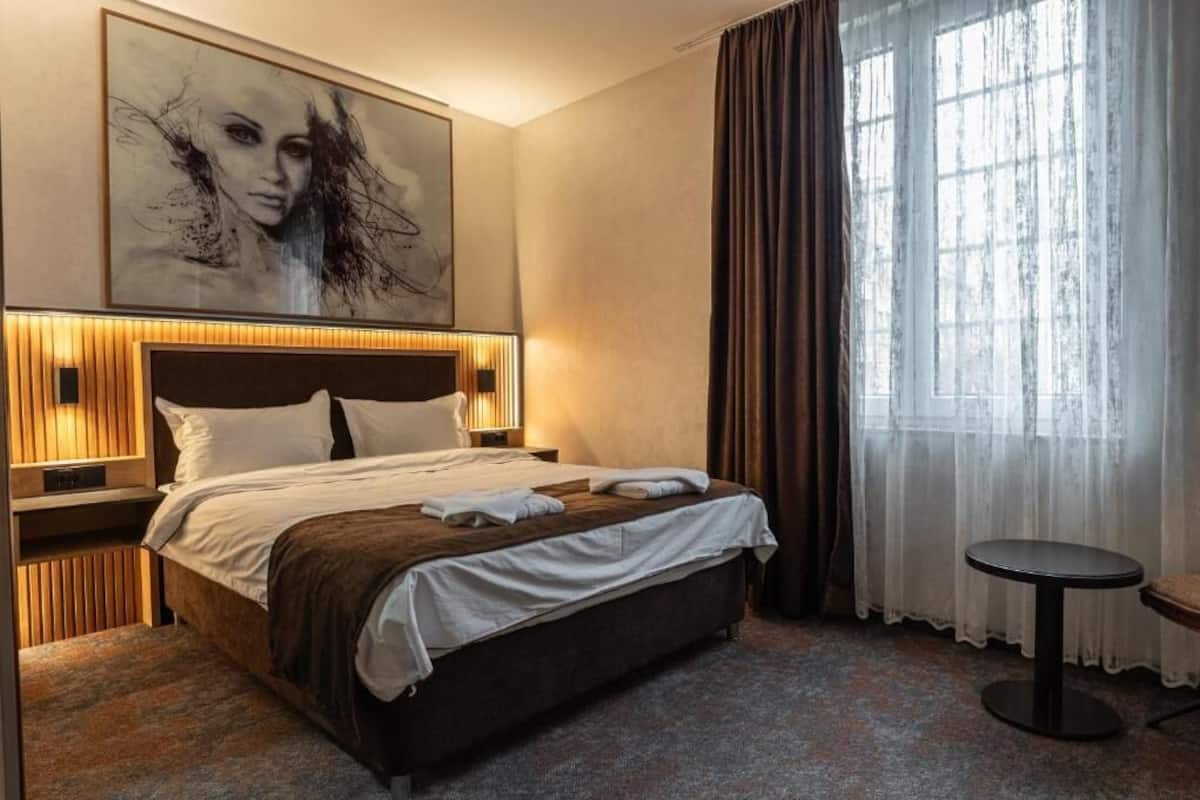 Una habitación de hotel con una cama grande, una mesita de noche, una silla, un ventanal con cortinas y un cuadro en la pared.