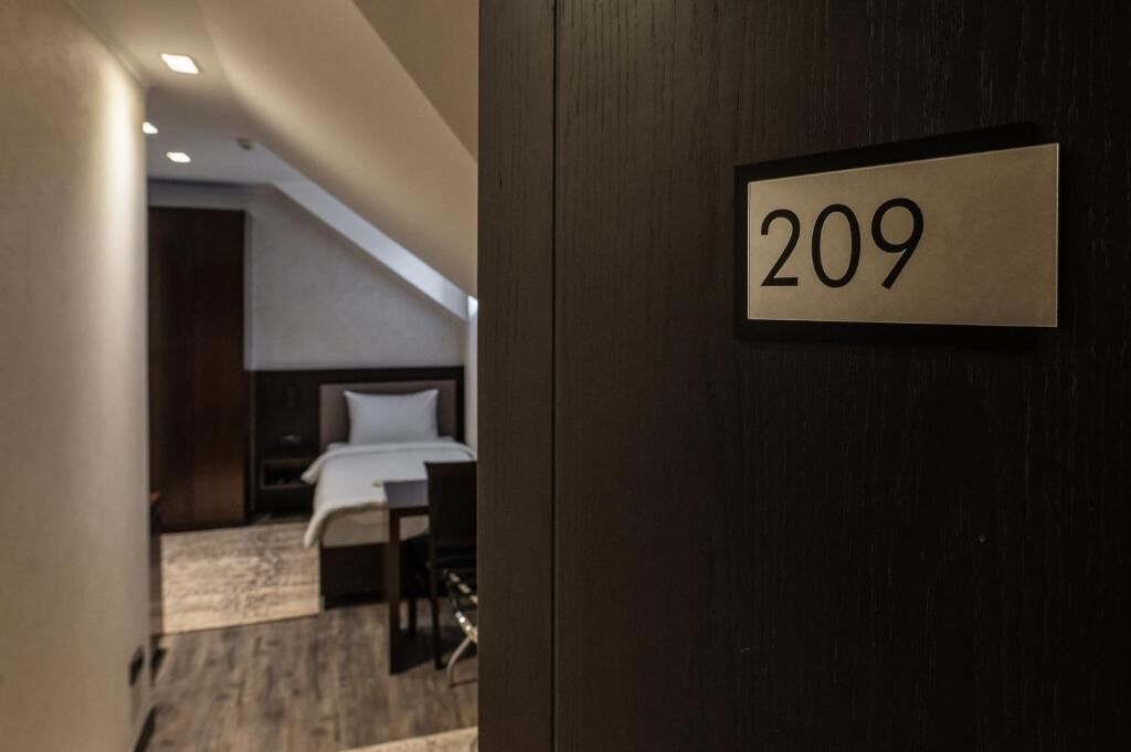Una camera d'albergo con un letto, una scrivania e una sedia. La porta, in legno, porta il numero 209.