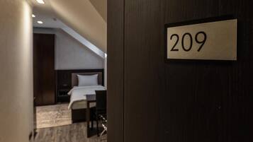 Una camera d'albergo con un letto, una scrivania e una sedia. La porta, in legno, porta il numero 209.