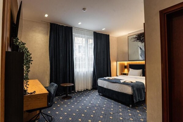 A hotel room with a bed, a desk, a chair, a small table, a potted plant, and a window with curtains. - Hotel Astral (Šabac)