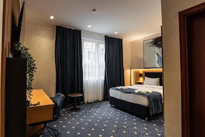 A hotel room with a bed, a desk, a chair, a small table, a potted plant, and a window with curtains.
