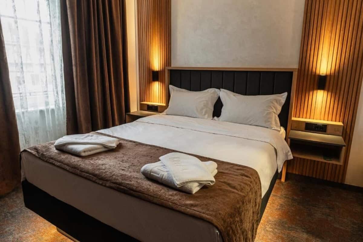 Habitación de hotel con una cama grande, dos mesitas de noche con lámparas, una ventana con cortinas y una alfombra marrón.