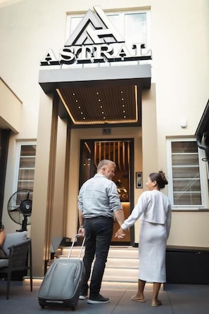 A hotel entrance with a sign reading 'ASTRAL,' a man holding a suitcase, and a woman standing beside him.
