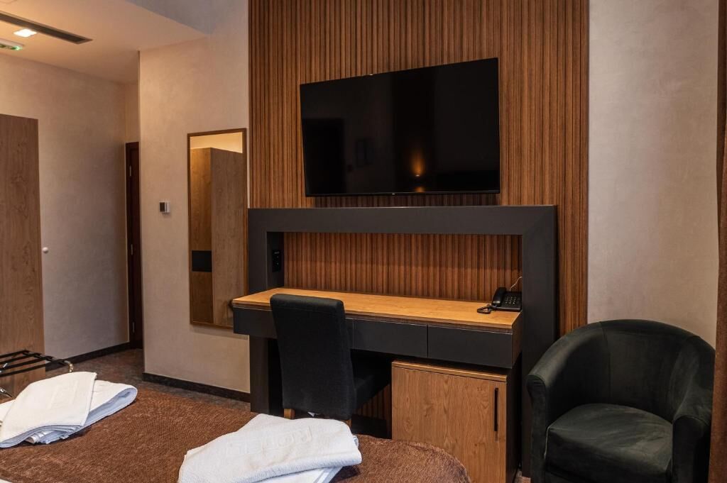 Een hotelkamer met een flatscreen televisie bevestigd aan een houten wandpaneel, een bureau met een stoel en een bed met een witte deken.