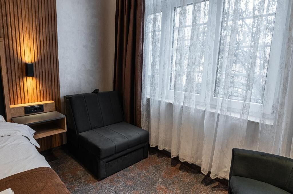 Ein Zimmer mit einem Bett, einem kleinen schwarzen Sofa, einer Wand aus Holzpaneelen mit einer Leuchte, einem Fenster mit durchscheinenden Vorhängen und Blick auf Bäume.
