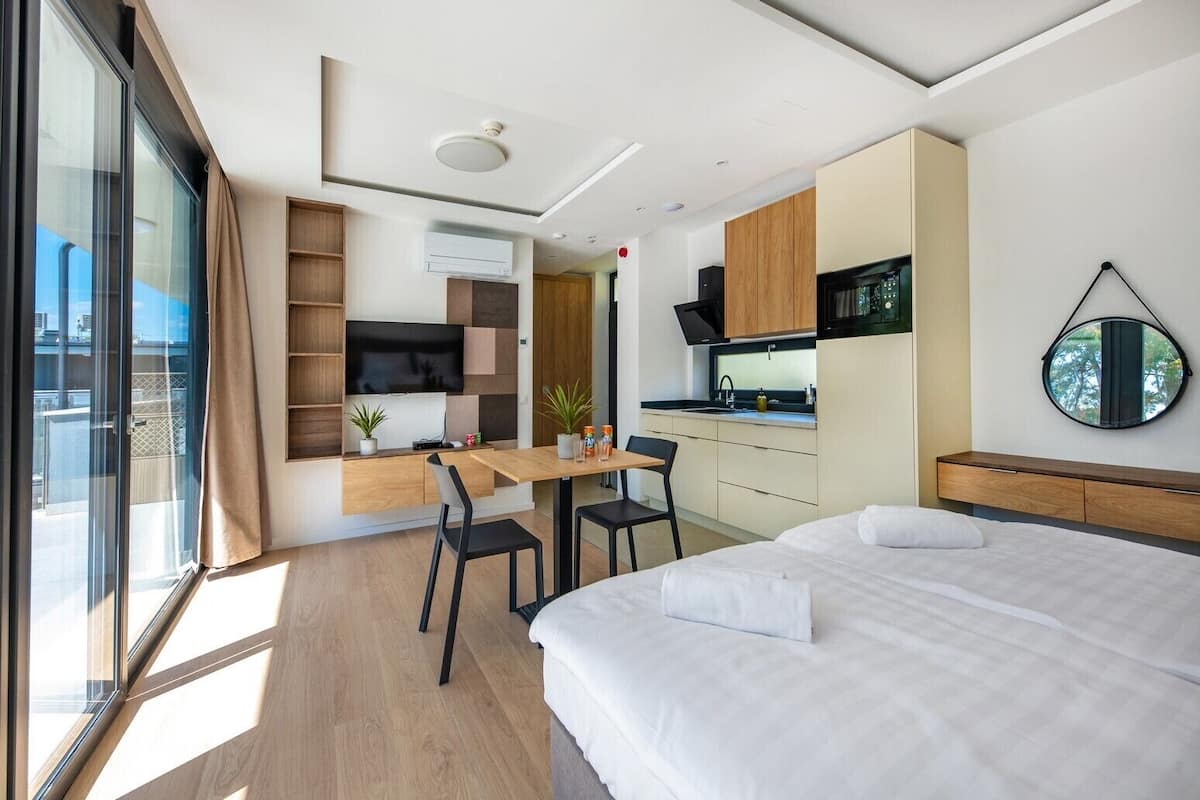 A modern hotel room with a bed, a desk, a kitchenette, and a large window.