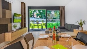 Un comedor moderno con una mesa de madera preparada para dos comensales, un sofá y vista a un jardín a través de un ventanal.