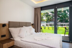 A modern hotel room with a large bed, a wooden nightstand, a built-in wall lamp, and a sliding glass door leading to a garden. - H55 Apartmanházak (Balatonlelle)