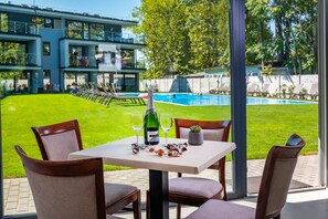 A table with a bottle and glasses, chairs, and a view of a swimming pool and building. - H55 Apartmanházak (Balatonlelle)