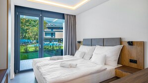 Una habitación de hotel moderna con una cama grande, cabecera de madera y vistas a una piscina y vegetación.