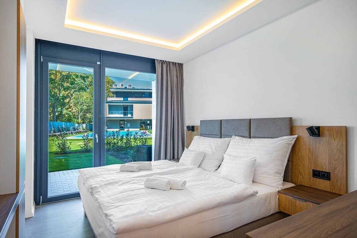 A modern hotel room with a large bed, a wooden headboard, and a view of a pool and greenery outside.
