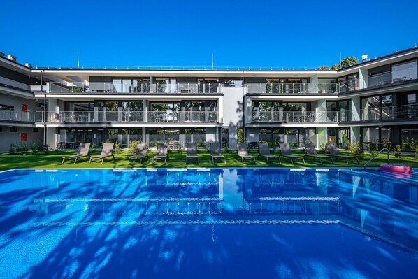 Ein modernes Hotel mit einem zentral gelegenen Schwimmbad, mehreren Balkonen und einer gepflegten Rasenfläche.