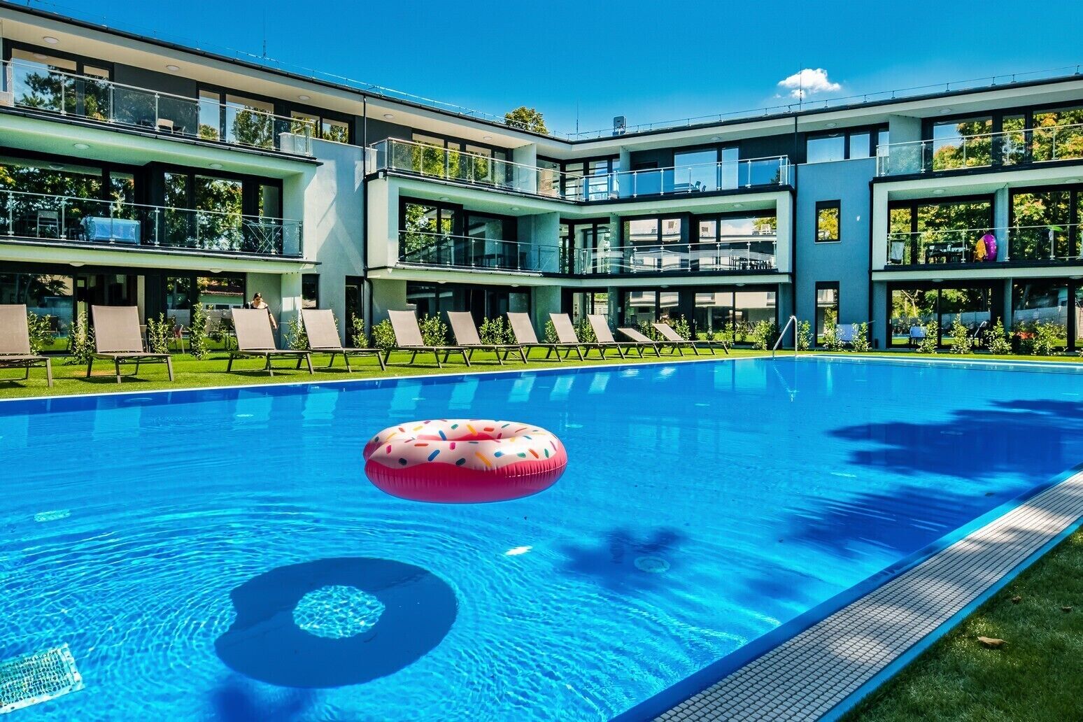 A modern hotel with a swimming pool, lounge chairs, and a clear blue sky.