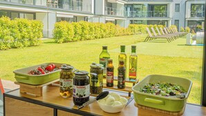 Una encimera de cocina con diversos alimentos y condimentos, un bol de huevos, y vistas a un edificio moderno con piscina y tumbonas.