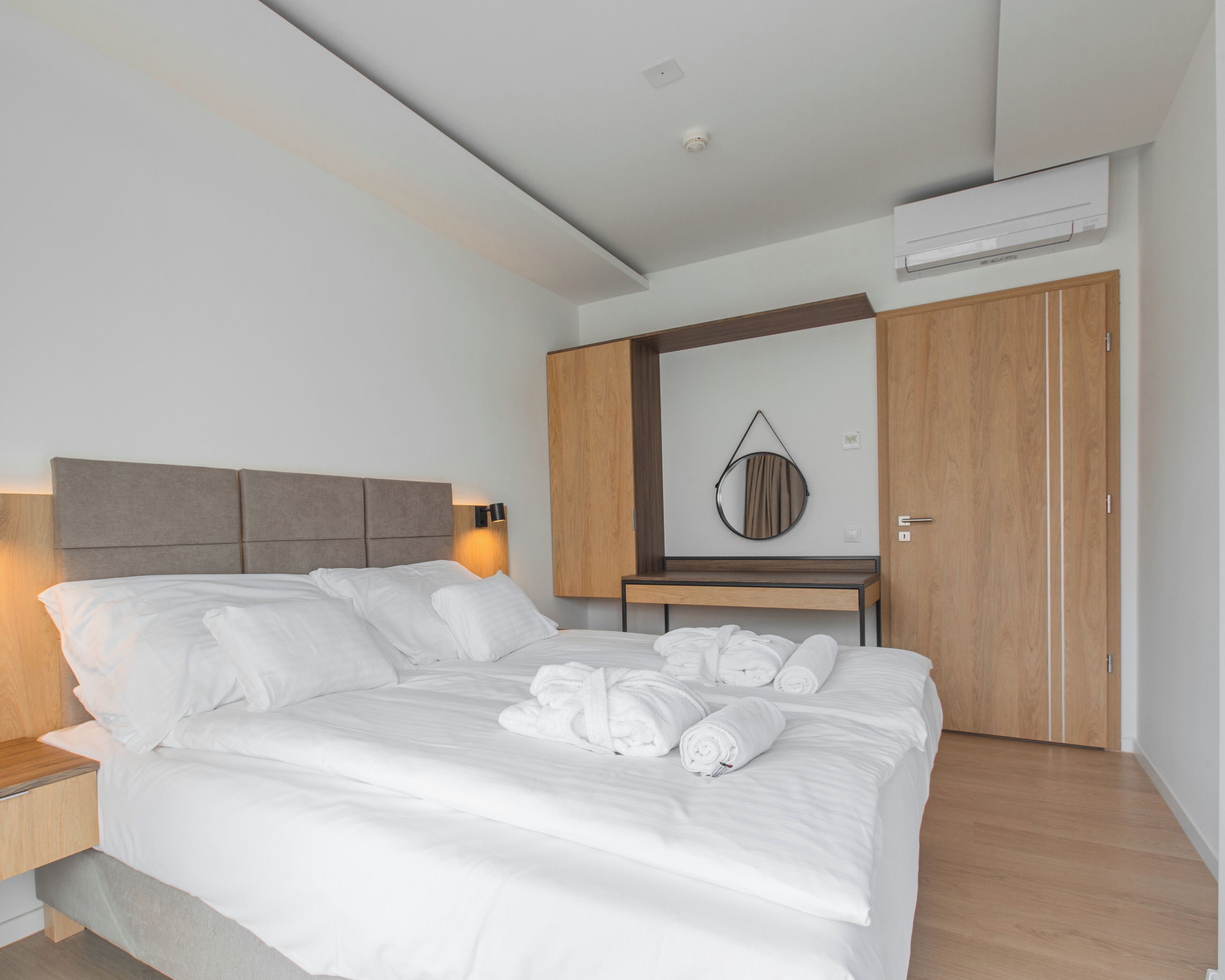 Una habitación de hotel moderna con una cama grande, un armario de madera, un espejo redondo y un equipo de aire acondicionado instalado en la pared.