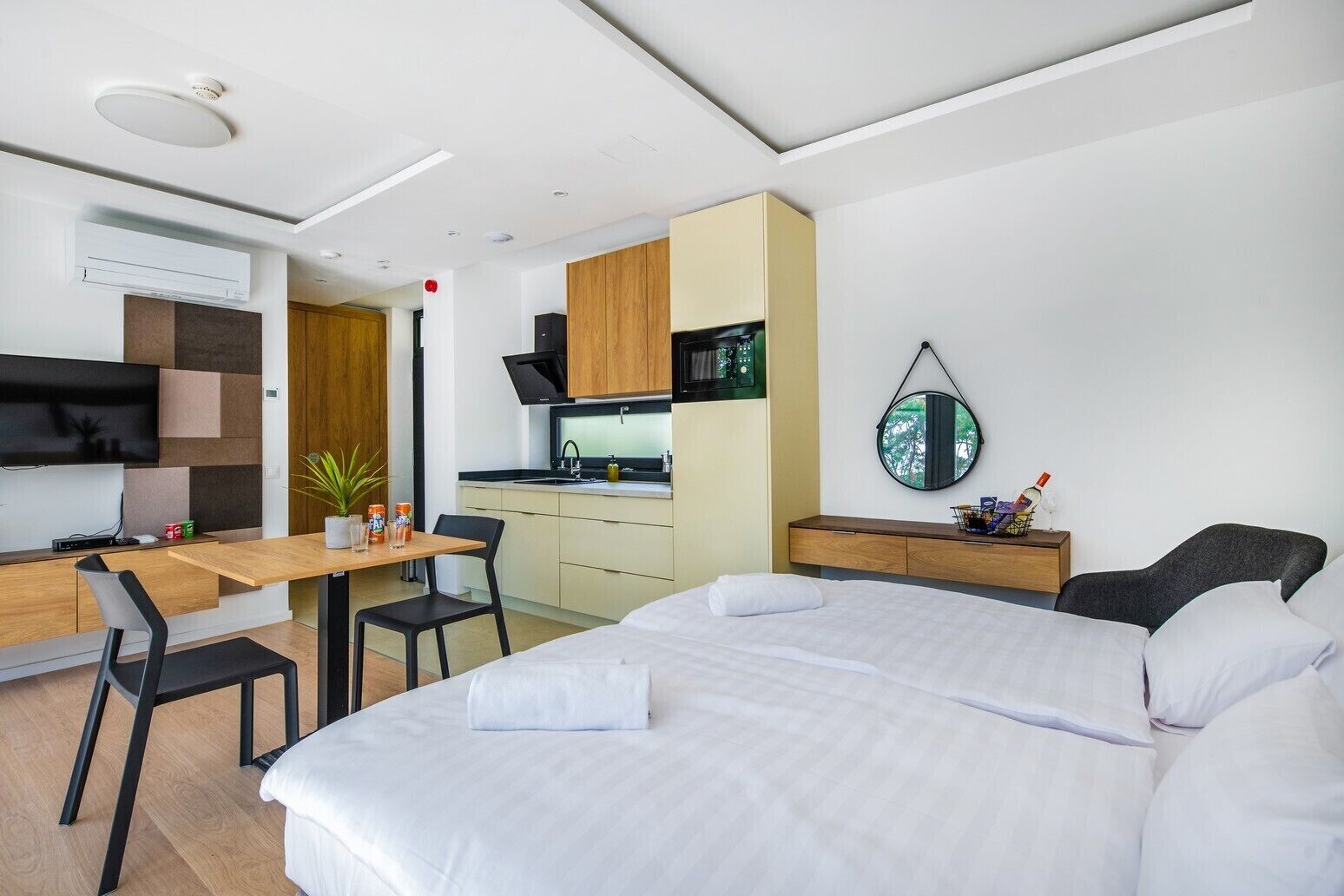 Quarto de hotel moderno com cama, escrivaninha com cadeira, cozinha compacta, televisão e espelho.