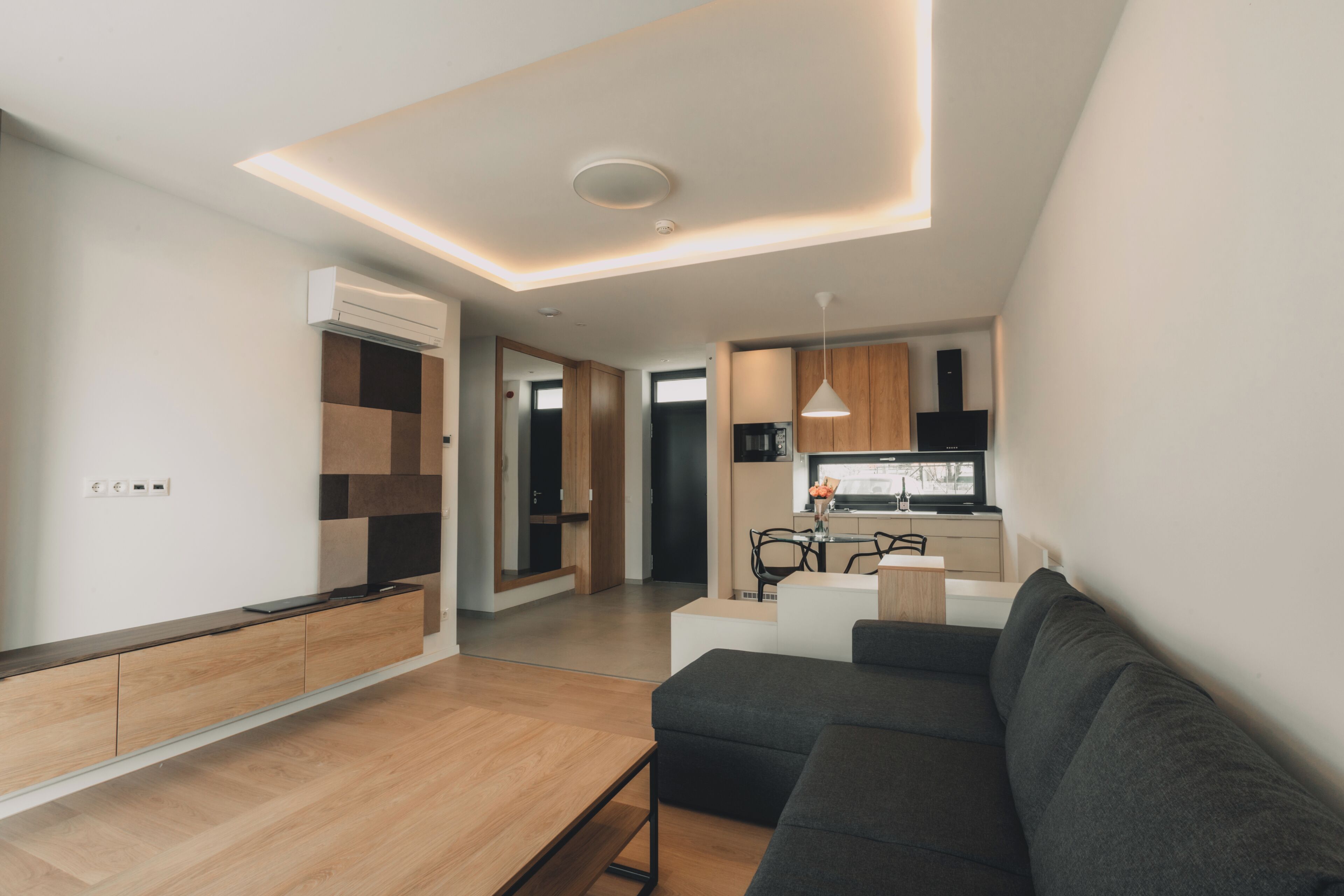 A modern living room with a sofa, a coffee table, and a TV. There is a kitchen area with cabinets and a dining table.