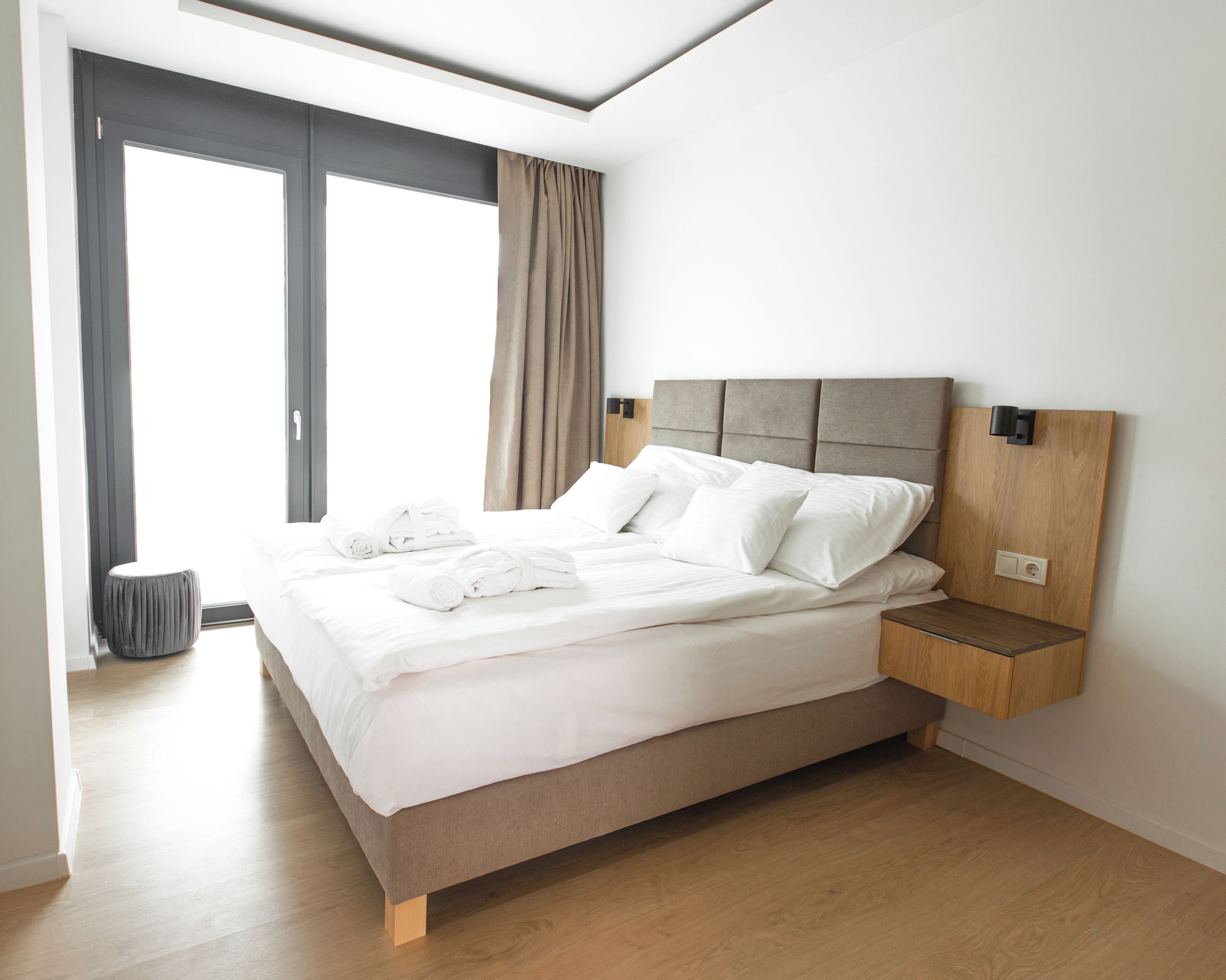 A modern hotel room with a large bed, a wooden nightstand, a grey ottoman, and a view of the outdoors.