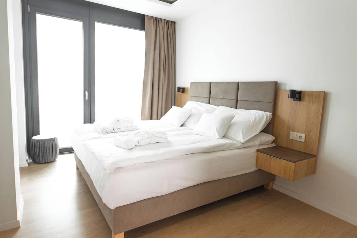 A modern hotel room with a large bed, a wooden nightstand, a grey ottoman, and a view of the outdoors.