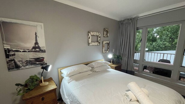 A bedroom with a bed, bedside lamps, a nightstand, a mirror, and a window with a view of greenery. - Room-luxury Stay in Pretoria! Fast Wi-fi (Pretoria)