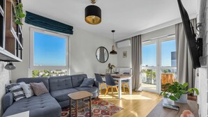 A modern living room with a grey sectional sofa, a small wooden table, and a dining area with a white table and chairs. - Praga Apt - 57m2, Parking, Balcony - by Rentujemy (Warszawa)