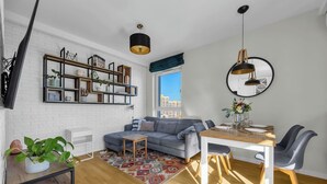 A modern living room with a grey sofa, wooden dining table, and a large window with a city view. - Praga Apt - 57m2, Parking, Balcony - by Rentujemy (Warszawa)