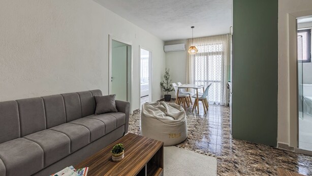 A living room with a sofa, a coffee table, a bean bag, a dining area with a table and chairs, and a kitchen visible through an open door.