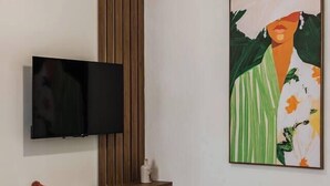 A modern living room with a flat-screen TV, wooden panel wall, a large abstract painting, and a coffee table with books and a plant.