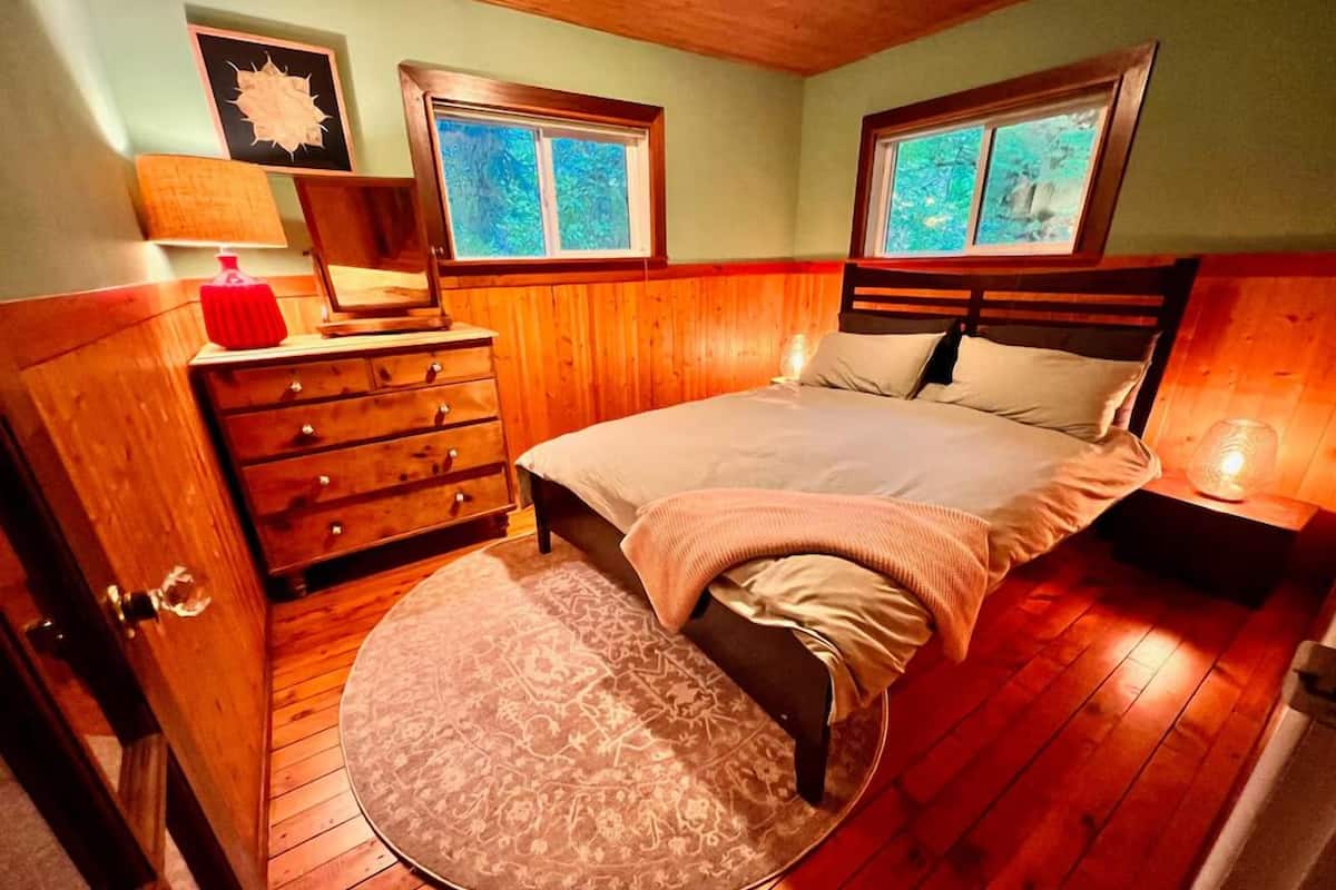 Un dormitorio con piso de madera, una cama con almohadas, un tocador, una lámpara y dos ventanas.