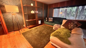 Een woonkamer met een bank, een vloerlamp, een boekenkast en een raam met uitzicht op bomen.