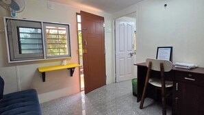 A room with a bed, a desk, a chair, a window, a door, and a fan. - Nirmala's Farm House (Courtallam)