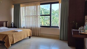 A hotel room with a bed, a bedside table, a window with curtains, and a view of greenery outside. - Nirmala's Farm House (Courtallam)