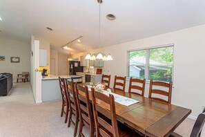 A dining area with a wooden table and chairs, a kitchen in the background, and a window with blinds.